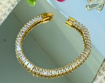Puede incluir: Un brazalete dorado con una fila de piedras transparentes en forma de baguette.