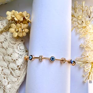 May include: A gold bracelet featuring blue evil eye charms and clear crystal accents. The bracelet is made from a thin, flexible cable chain.
