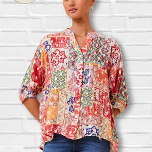 May include: A vibrant patchwork blouse with a V-neck and short sleeves. The blouse showcases a mix of red, yellow, blue, and green patterns. The La Capa Moda Italiana logo is visible. The model is wearing blue jeans.