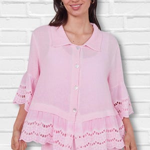 May include: A light pink button-down shirt featuring a classic collar, three-quarter length sleeves with ruffled cuffs, and a ruffled hemline. Both the cuffs and the hem are detailed with intricate white eyelet embroidery and scalloped edges.