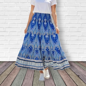 May include: A woman wearing a white t-shirt and a long, tiered blue skirt with a paisley pattern. The skirt has a detailed border design. The image includes a logo with text that reads "LaCapa Moda Italiana".
