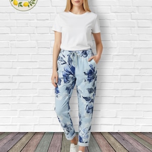 May include: A woman in a white t-shirt and light blue floral print trousers. The trousers have a drawstring waist and are cuffed at the ankles. She wears white trainers. The background is a white brick wall and a wooden floor.