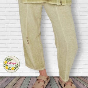 May include: Light green linen pants with three buttons on the side, paired with beige sandals. The pants have a relaxed fit and are part of a casual outfit. The LaCapa Moda Italiana logo is visible.