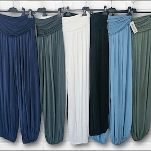 May include: A collection of six pairs of loose-fitting trousers hanging on black plastic hangers. The trousers feature a wide, gathered, fold-over waistband and tapered legs with elasticated cuffs. The colours include navy, dark teal, white, black, light blue, and sage green.