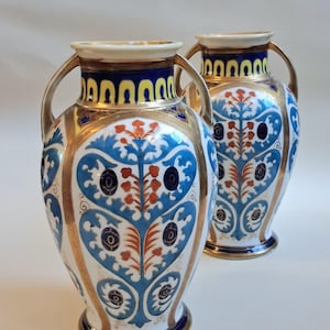 Puede incluir: Dos jarrones decorativos de cerámica con asas y adornos dorados. Los jarrones presentan una base blanca con motivos florales azules y naranjas. Los bordes son azules con detalles dorados. Un artículo de decoración del hogar clásico.