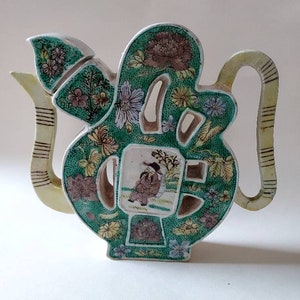 Könnte beinhalten: Eine grüne und gelbe Teekanne aus Keramik mit einem Blumendesign und einer gemalten Figur einer Person auf der Vorderseite. Die Teekanne hat eine einzigartige Form und ist ein dekoratives Stück.