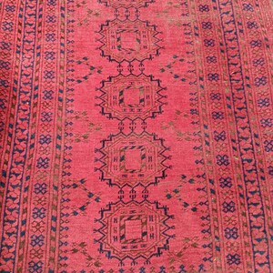 May include: A red and brown patterned rug with a central design of repeating geometric shapes. The rug has a border with a repeating pattern of small floral and geometric elements. The rug's color palette includes shades of red, brown, and blue.