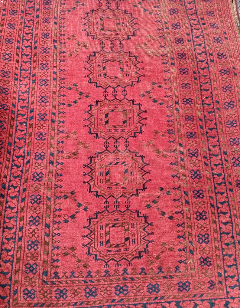 May include: A red and brown patterned rug with a central design of repeating geometric shapes. The rug has a border with a repeating pattern of small floral and geometric elements. The rug's color palette includes shades of red, brown, and blue.