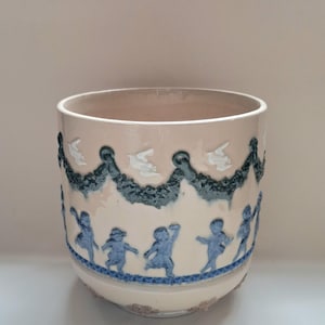 May include: A cylindrical ceramic planter with a cream-coloured base. The exterior features raised relief decorations in blue and white, including a series of dancing figures around the bottom and a garland pattern with white birds near the top rim.