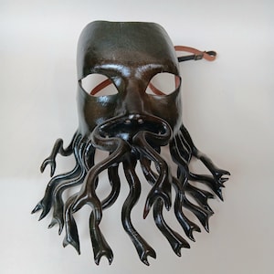 Op de afbeelding: Een donkergroen leren masker met een menselijk gezicht en een lange, stromende baard gemaakt van vele ranken. Het masker heeft twee ooggaten en een kleine neus.