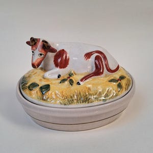 May include: A ceramic butter dish featuring a white cow with brown spots and accents, resting on a bed of yellow hay. The dish has a light pink base with a matching lid, creating a charming farm animal theme.