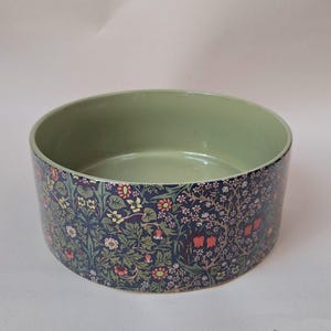 Puede incluir: Un cuenco redondo de cerámica con un estampado floral. El exterior presenta un fondo azul oscuro con un patrón repetido de flores y hojas en tonos rojos, rosas, amarillos y verdes. El interior es de color verde claro liso.
