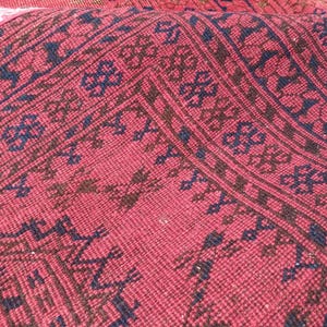 May include: Close-up of a section of a red rug with a detailed pattern. The rug features a repeating design in dark blue and brown, creating a textured appearance. The rug's fibers are visible, adding depth to the overall aesthetic.