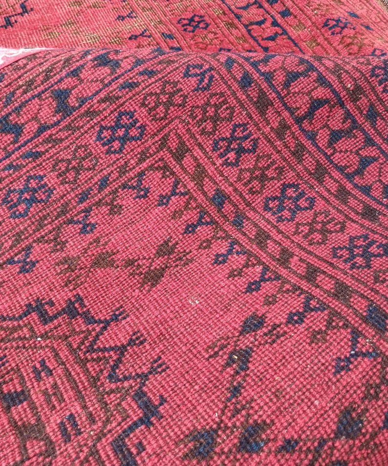 May include: Close-up of a section of a red rug with a detailed pattern. The rug features a repeating design in dark blue and brown, creating a textured appearance. The rug's fibers are visible, adding depth to the overall aesthetic.