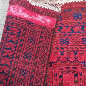 May include: A section of a red rug with intricate geometric patterns in navy blue and brown. The rug has a fringed edge and a folded section revealing the underside. The rug's design includes repeating floral and geometric motifs.