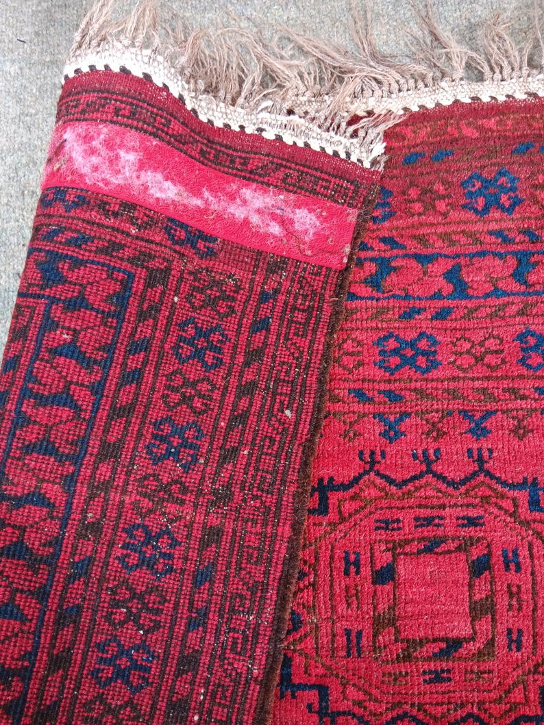 May include: A section of a red rug with intricate geometric patterns in navy blue and brown. The rug has a fringed edge and a folded section revealing the underside. The rug's design includes repeating floral and geometric motifs.