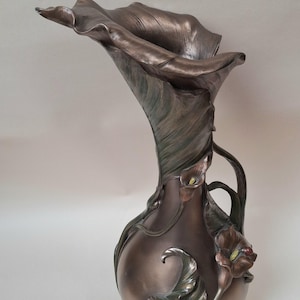 May include: A metallic bronze-toned vase shaped like a large calla lily flower. The vessel features a wide, flared rim, a bulbous base, and sculpted leaf and smaller flower accents along the exterior, finished with subtle green and brown hues.
