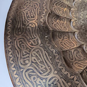 May include: Close-up of a brass decorative plate with intricate etched patterns. The plate features a detailed design of swirling lines and shapes, creating a textured surface. The plate's edge has a decorative border.