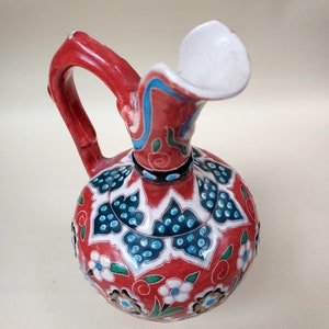 May include: A red ceramic jug with a white spout and a floral design in blue, green, and white.