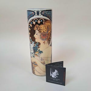 May include: A cylindrical ceramic vase featuring a colourful Art Nouveau illustration of a woman in profile holding a feather. Beside the vase stands a small black informational card with a portrait of Alphonse Mucha and the text The Feather 1899.