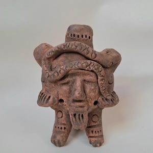 以下が含まれることがあります： 茶色のテラコッタ彫刻で、目を閉じて髭を生やした人間のような顔が中央にあり、その周りを蛇が取り巻いています。このフィギュアには、円形や線状の彫り込みが施された2本の脚と、小さな穴が開いた円筒形の頂部があります。
