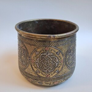 May include: A cylindrical metal container with an aged, antique appearance. The container is primarily gold-toned with intricate etched patterns and Arabic script. A central circular design features a complex knot pattern with hints of pink and silver.