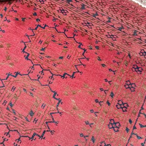 May include: A close-up of a red rug with intricate geometric patterns in navy blue and brown. The rug has a textured surface and a fringe along one edge. The design includes repeating floral and geometric motifs.
