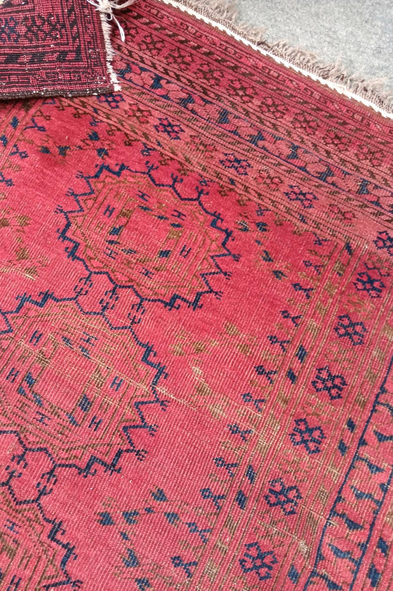May include: A close-up of a red rug with intricate geometric patterns in navy blue and brown. The rug has a textured surface and a fringe along one edge. The design includes repeating floral and geometric motifs.