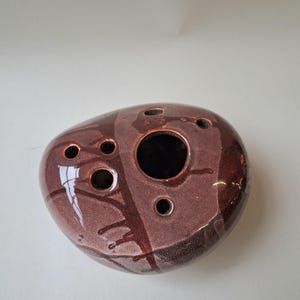 Könnte beinhalten: Braune Keramik-Okarina mit gesprenkelter, glänzender Oberfläche. Das Instrument hat mehrere kreisförmige Löcher unterschiedlicher Größe, darunter eine große zentrale Öffnung. Dunklere braune Streifen sorgen für visuelles Interesse.