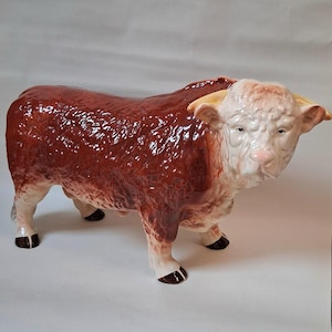 Puede incluir: Figura de toro de cerámica con acabado brillante. El toro tiene un cuerpo marrón rojizo, cara blanca y cuernos color canela. Las pezuñas son negras. La figura está de pie sobre sus cuatro patas.