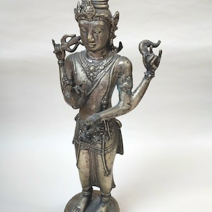 Puede incluir: Una estatua de metal de color plateado de una deidad con cuatro brazos, que lleva una corona y joyas elaboradas. La estatua está de pie sobre una base en forma de loto.