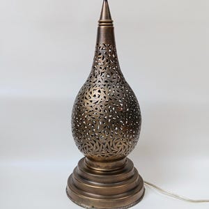 May include: Bronze-coloured table lamp with a decorative, pierced metal shade. The lamp has a rounded base and a tall, slender body with intricate cut-out patterns. The lamp is approximately 51 cm tall.