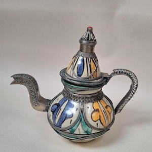 May include: A ceramic teapot featuring a cream-coloured base with hand-painted blue, yellow, and green floral motifs. The teapot is encased in intricate silver-toned metal filigree work on the handle, spout, and lid, topped with a small red bead.