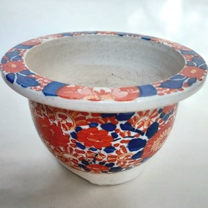 Peut inclure: Un pot en céramique blanche avec un motif floral rouge, bleu et orange. Le pot a un bord large et un fond arrondi.