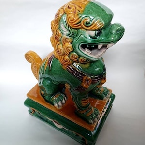 Pode incluir: Uma estátua de cão foo de cerâmica verde e dourada com uma expressão feroz. A estátua está em pé sobre uma base quadrada com detalhes verdes e dourados.