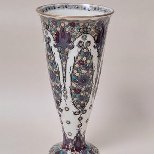 May include: A tall, flared ceramic vase with a white base featuring intricate, repeating patterns in shades of purple, teal, and gold. The design includes stylized floral motifs and small circular dots arranged in vertical panels, finished with a gold rim.