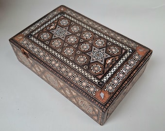May include: A rectangular wooden box with intricate inlaid mosaic designs. The box features a repeating pattern of stars and geometric shapes in shades of brown, white, and black. The box is closed and appears to be a decorative storage item.