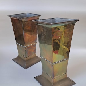 May include: A pair of square-shaped metal vases with a polished, iridescent bronze and gold finish. Each vase features a flared top, a tapered body, and a decorative twisted rope-like band encircling the middle section, resting on a square base.