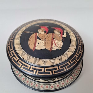 May include: A round, black decorative box with a lid featuring a Greek warrior design. The lid has a black background with two warriors, shields, and spears. The box has a geometric border in gold and cream.