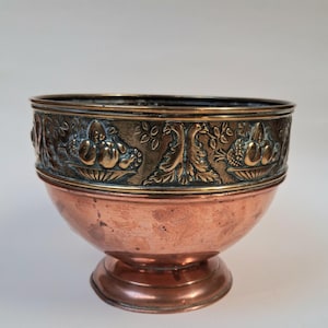 May include: A copper bowl with a pedestal base. The upper rim features a detailed relief design with fruit and foliage. The bowl has a warm, reddish-brown color, with a contrasting darker rim. The bowl is likely vintage and decorative.
