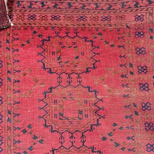 May include: A close-up of a red rug with intricate geometric patterns in navy blue and brown. The rug has a textured surface and a fringed edge. The design features repeating shapes and borders.