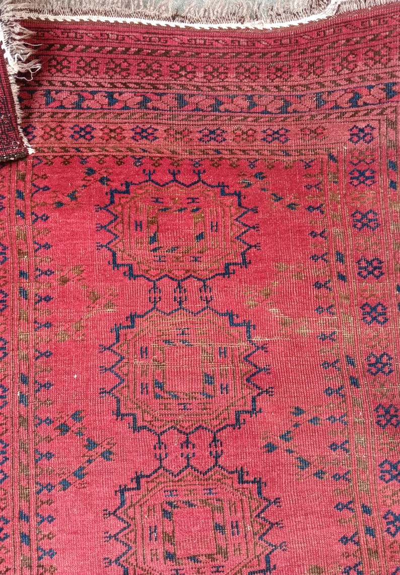 May include: A close-up of a red rug with intricate geometric patterns in navy blue and brown. The rug has a textured surface and a fringed edge. The design features repeating shapes and borders.