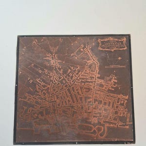 May include: A square copper printing plate featuring an engraved map of Liverpool. The metallic surface displays intricate street layouts, a compass rose, and a decorative cartouche in the upper right corner containing the text Liverpool.