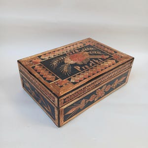 May include: A wooden box with a black background and intricate straw inlay designs. The lid features a wheat sheaf design with the word "Prosit" in the center.