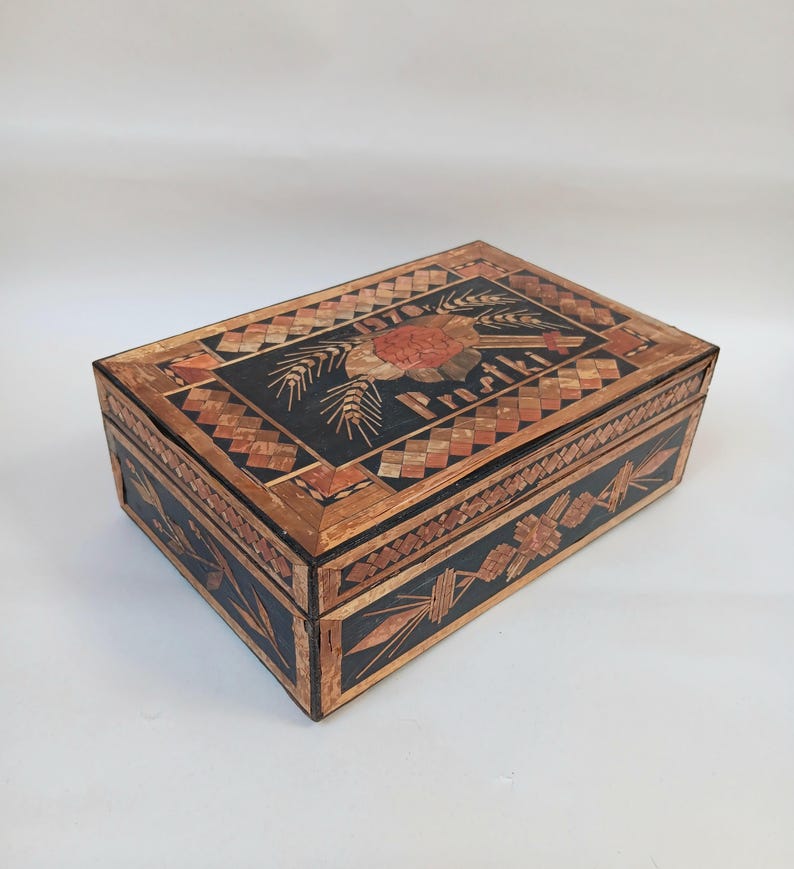 May include: A wooden box with a black background and intricate straw inlay designs. The lid features a wheat sheaf design with the word "Prosit" in the center.