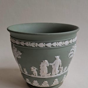 May include: A sage green ceramic planter with white bas-relief classical figures and foliage. The planter has a wide rim and a decorative band of white leaves. The detailed relief depicts people and animals in a pastoral scene.