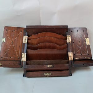 Könnte beinhalten: Ein dunkelbrauner Schreibtisch-Organizer aus Holz mit zwei aufklappbaren Seitenteilen, die mit eingelegten Schnurmustern und Messingbeschlägen versehen sind. Der Mittelteil enthält vier gestufte, geschwungene Trennwände und eine Schublade mit Messingverschluss.