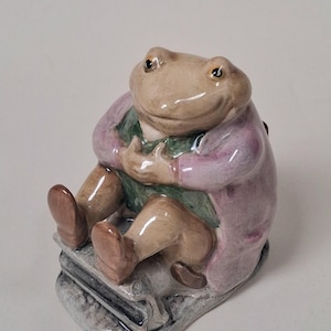 Könnte beinhalten: Eine Keramikfigur eines sitzenden Frosches, der einen hellvioletten Mantel über einer grünen Weste trägt. Der Frosch hat braune Haut, dunkle Augen und sitzt auf einem grauen, rechteckigen Sockel. Die Oberfläche der Figur hat ein glänzendes Finish.