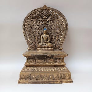 May include: A gold-colored metal statue of a seated Buddha with a decorative, ornate base and a detailed, intricate halo. The statue is on a white background.