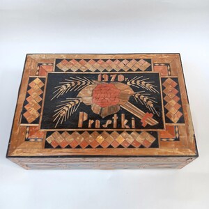 May include: A wooden box with a black background and a red flower design. The box is decorated with a geometric pattern of wood pieces. The text "1970r. Prustki" is visible on the box.